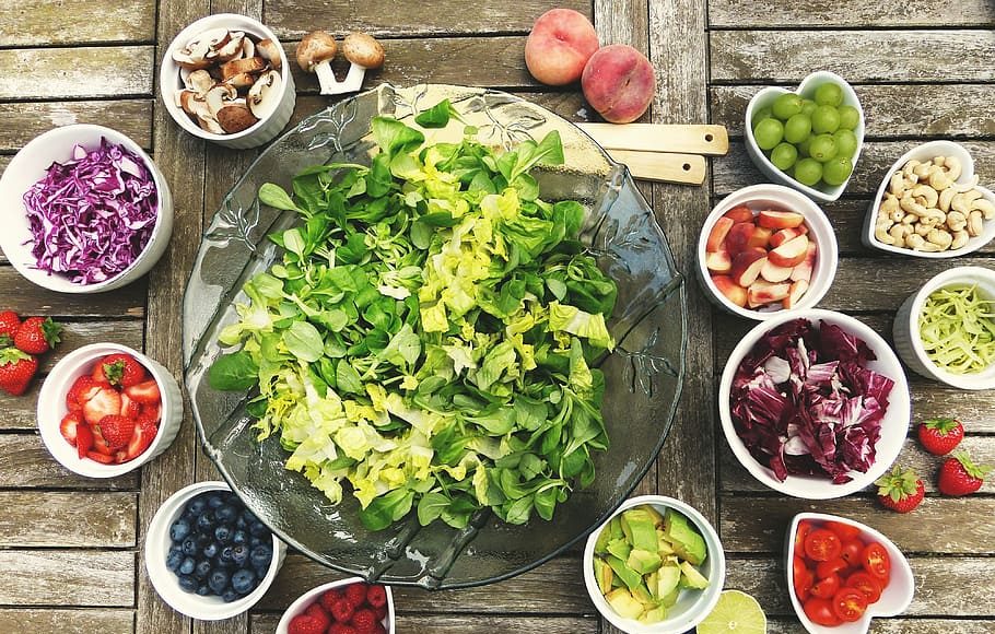 Prevent Chronic disease by adding more healthy food to your diet like a large glass bowl of green salad, surrounded by smaller bowls of different fruits, nuts, and vegetables, on top of a wooden plank table.