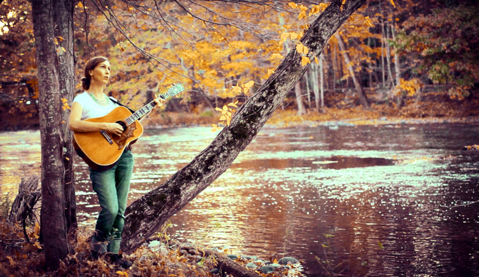 Katy Mantyk singing and playing guitar by a river.